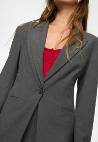 Blazer gris sur mesure avec une fermeture à un bouton, des revers crantés et deux poches avant. Porté sur un haut rouge boutonné.