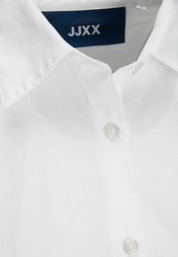 White button-up shirt with a classic collar, featuring a single round button and a woven label with "JJXX" in blue at the neckline.