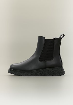 Bottine Chelsea noire avec semelle texturée, panneaux latéraux élastiques et languette de traction sur fond clair neutre.