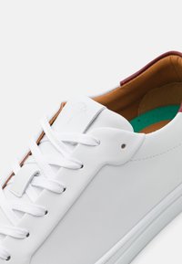 Vit lädersneaker med slät yta, platta snören och brunt läderinteriör. Har en subtil präglad logotyp och en grön sula.