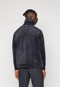 Jachetă pulover din fleece navy cu guler înalt, textură netedă și detaliu logo pe spate; prezintă buzunare laterale și o croială lejeră.