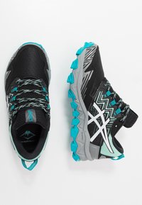 Baskets de sport noires avec des accents turquoise, dotées d'une tige en maille texturée, d'un motif en zigzag et d'une semelle grise robuste avec des pointes pour une meilleure adhérence.