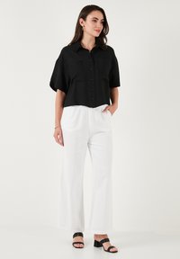 Camicetta nera corta con maniche corte e taschini frontali, abbinata a pantaloni bianchi a gamba larga e sandali col tacco neri. Texture liscia su tutta la superficie.