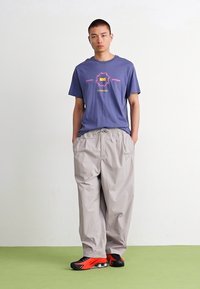 Grå, løstsiddende bukser i kombination med en lilla t-shirt med et iøjnefaldende, multicolor grafisk design. Sorte og røde sneakers fuldender outfittet.