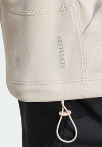 Top atletico beige chiaro con una trama liscia, caratterizzato dal marchio AEROREADY e un dettaglio con cordino all'orlo. Cuciture piatte visibili.