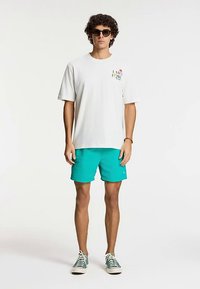 Bijela majica kratkih rukava s šarenim printom na prsima, uparena s tirkiznim kratkim hlačama i zelenim tenisicama s uzorkom.