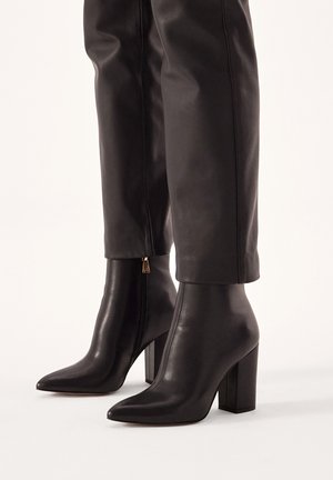 Bottines en cuir noir avec des bouts pointus, des talons larges et des fermetures éclair sur le côté. Texture lisse avec un design élégant et moderne.