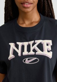 Zwarte katoenen T-shirt met een groot, gestileerd "NIKE"-logo in roomwit en paars, met daaronder een klein wit en paars Nike swoosh-logo.