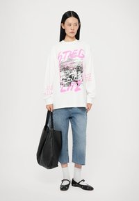 White long-sleeve t-shirt with pink graphic and text, paired with light blue cropped jeans, black handbag, white socks, and black shoes.