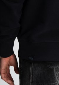 Marineblauwe sweatshirt met ribboorden aan de mouwen en onderkant, gemaakt van zachte stof. Heeft een klein label aan de onderzijde met de tekst "Butcher of Blue".