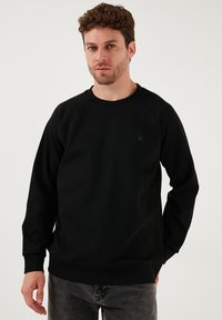 Črna trenerka s crewneck izrezom in dolgimi raglan rokavi, izdelana iz mehkega materiala; vsebuje majhen logotip na prsih in rebraste manšete.