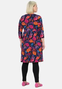 Kukallinen mekko, jossa on kirkkaat kukat oranssina, vaaleanpunaisena ja sinisenä violetilla taustalla. Kolmannen neljänneksen hihat, polvimittainen leikkaus, yhdistetty mustiin legginsseihin.