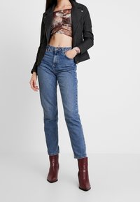 Sort faux læder motorcykeljakke, kort mønster top, højtaljede blå jeans og brune spidse ankelstøvler med en tekstureret overflade.