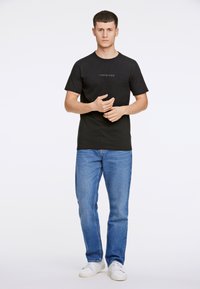 Musta puuvillapaita, jossa on pyöreä kaula-aukko ja rintataskussa 'LINDBERGH'-logo, yhdistettynä vaaleansinisiin farkkuihin ja valkoisiin tenniskenkiin.