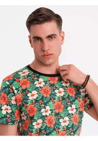 Bloemrijke t-shirt met korte mouwen op een groene basis, met rode en witte hibiscuspaterns. Zwarte ronde halslijn en zachte textuur.