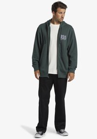 Sudadera con cremallera de color verde oscuro, con bolsillos tipo canguro, llevada sobre una camiseta blanca, combinada con pantalones negros y zapatillas negras.