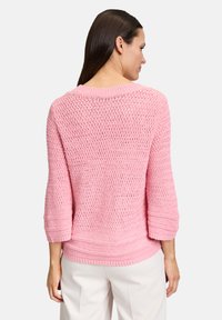 Maglione rosa lavorato a maglia con un design a trama aperta e ampia, maniche a tre quarti e orlo a costine. La texture sembra leggera e traspirante.