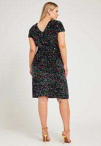 Abito nero con paillettes multicolori, maniche a cappuccio e design con scollo a V, finitura texturizzata. Indossato con sandali dorati con tacco alto.