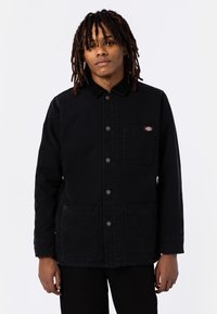 Veste en toile noire avec un col, des boutons-pression, des poches avant et un petit patch logo sur la poitrine gauche. Design minimaliste.