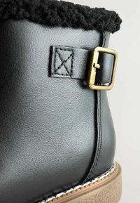 Schwarze Lederstiefel mit einem flauschigen schwarzen Kragen, verzierten Messing-Schnallen, strukturiertem Obermaterial und einer hellbraunen Sohle mit genähter Kante.