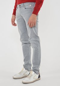 Person in hellgrauen Jeans, weißen Sneakers mit beige und schwarzen Details und einem roten Langarmoberteil, steht vor einem weißen Hintergrund.