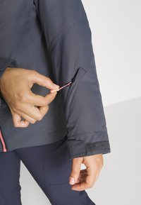 Ciemnoszara kurtka z kontrastowym różowym zamkiem, wyposażona w kieszeń zapinaną na zamek błyskawiczny na lewym rękawie oraz regulowane mankiety na rzep. Gładka tekstura.