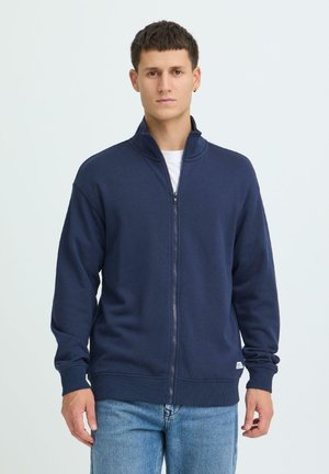 Marineblauwe sweatshirt met ritssluiting, hoge kraag, geribde manchetten en zoom, zachte textuur, gedragen over een wit T-shirt, gecombineerd met lichtblauwe spijkerbroek.