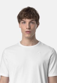 T-shirt bianco in cotone con scollo rotondo e maniche corte, caratterizzato da un piccolo logo tono su tono sul petto sinistro. Texture liscia, vestibilità casual.