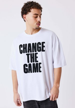 Jonge man met krullend haar, droeg een wit oversized T-shirt met gedurfde zwarte tekst "CHANGE THE GAME," kijkt naar beneden en opzij.
