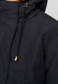 Chaqueta con capucha negra de tejido texturizado, que presenta un cordón con un tope de metal y un sutil detalle en las costuras.