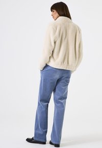 Frau steht mit Händen in den Taschen, trägt eine cremefarbene Fleecejacke, hellblaue Cordhosen und schwarze Slipper vor weißem Hintergrund.