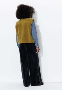Gilet en polaire vert olive, avec des manches longues en denim en dessous, associé à un pantalon large noir. Tissu texturé avec une coupe décontractée.