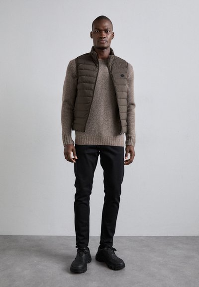 Gevoerde olijfgroene vest over een beige trui, gecombineerd met zwarte slim-fit jeans en zwarte chunky sneakers. Glad materiaal en geribde manchetten.
