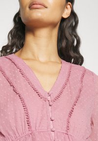 Robe rose texturée avec un décolleté en V, de délicates fermetures à boutons et un ourlet à volants le long du décolleté et des manches.