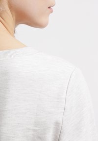 Boční profil osoby v svetlu šedém pleteném svetru, ukazující rameno a krk na jednobarevném pozadí.