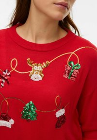 Maglione rosso con disegni natalizi in paillettes dorate: una renna, regali, un albero di Natale e calze, con tessuto lavorato a maglia strutturato.