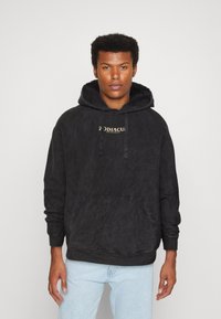Schwarzer Kapuzenpullover aus weichem Stoff, mit einer vorderen Kängurutasche und einem goldenen "Zodiac"-Logo auf der Brust. Lockere Passform, strukturierte Optik.