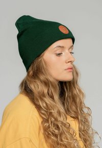 Zielona, prążkowana czapka z dzianiny z okrągłą, brązową skórzaną metką z przodu. Czapka ma luźny krój i lekko spiczasty szczyt.