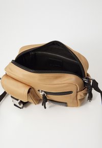 Tan shoulder bag made of smooth synthetic material; features black zippers, a front pocket, and an adjustable strap. Interior is black fabric.