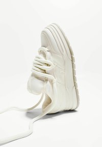Chaussure de sport blanche avec une surface texturée, un bout rond, des lacets ronds et une semelle épaisse et striée pour un meilleur adhérence et confort.