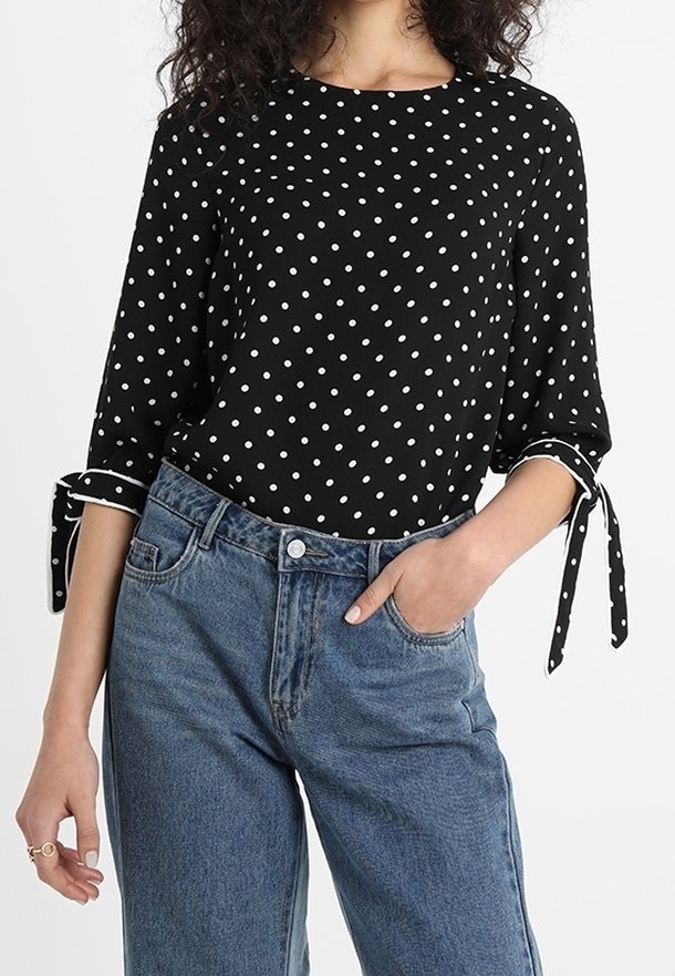 Černá halenka s puntíky, tříčtvrtečními rukávy a zavazovacími manžetami, v kombinaci s modrými džínami s vysokým pasem. Hladká tkanina, kulatý výstřih.