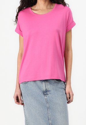 Femme portant un t-shirt rose vif à manches courtes et une jupe en jean bleu clair, debout sur un fond blanc.
