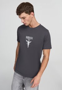 T-shirt grigia a maniche corte con una stampa grafica bianca di "NIRVANA LIVE '93" raffigurante una figura angelica con ali. Materiale in cotone morbido.