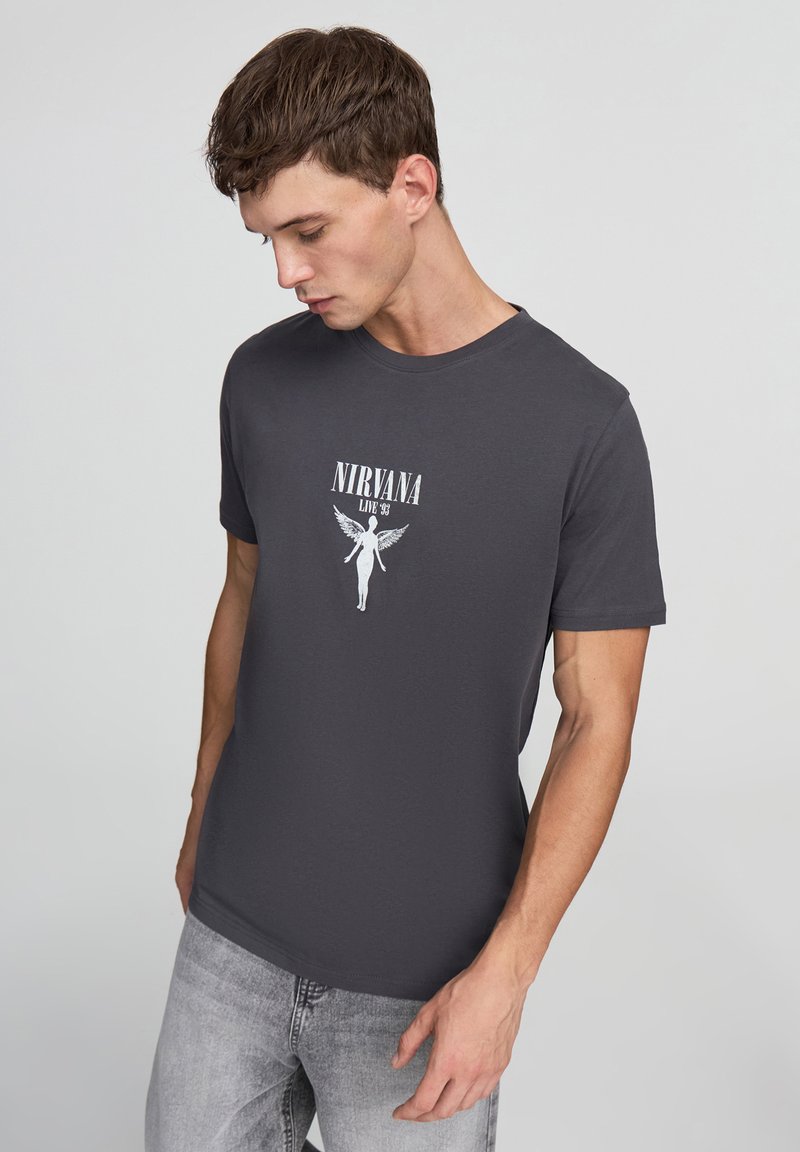 T-shirt grigia a maniche corte con una stampa grafica bianca di "NIRVANA LIVE '93" raffigurante una figura angelica con ali. Materiale in cotone morbido.