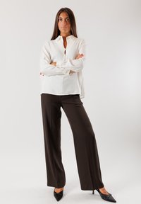 Camicia bianca con colletto e dettaglio a apertura a chiave abbinata a pantaloni larghi marroni e décolleté neri a punta su uno sfondo bianco.