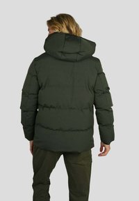 Personne aux cheveux blonds portant une doudoune vert foncé avec capuche, se tenant de dos contre un fond uni.