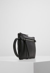 Musta nahkainen laukku, jossa on vetoketjullinen tasku, litteä suorakaiteen muoto, yläkahva ja hienovarainen tekstuuri. Sisältää sivulla roikkuvan hapsudetailin.