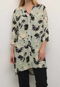 Tunique florale de couleur vert clair avec des fleurs noires et beiges, devant boutonné, col en V et manches trois quarts, fabriquée en tissu texturé.