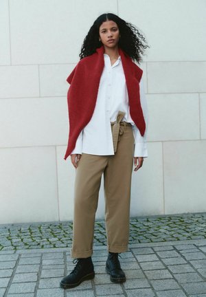 Femeie cu păr creț purtând un șal roșu, cămașă albă cu nasturi, pantaloni bej cu talie legată și cizme negre cu șiret, stând pe paviment de piatră.