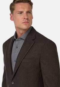 Brązowa blazer o fakturze wykonana z tkanej tkaniny, z pojedynczym guzikiem, klapami i kieszenią na piersi na szarym koszuli.